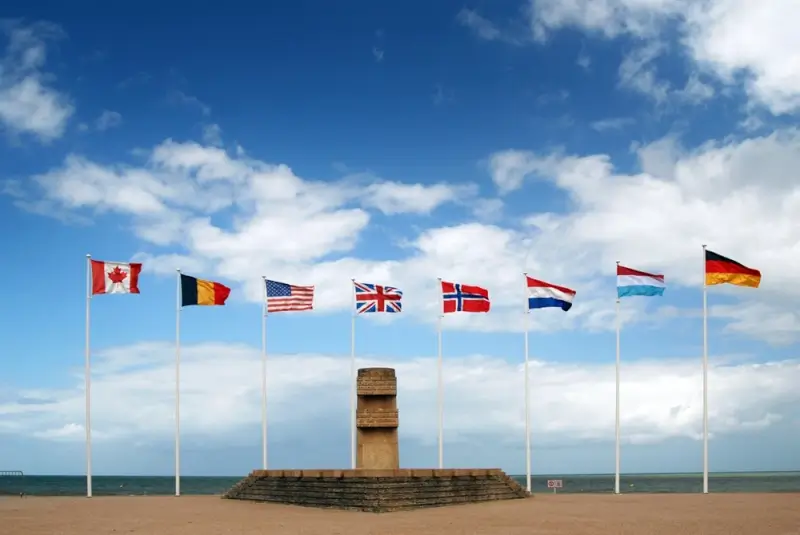 Allied nations flags flying above D-Day memorial monument on Normandy beach, honoring international cooperation during Operation Overlord