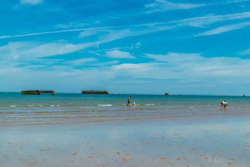 Remnants of Mulberry Harbour visible at low tide on Gold Beach Arromanches, with visitors exploring the historic D-Day artificial harbor