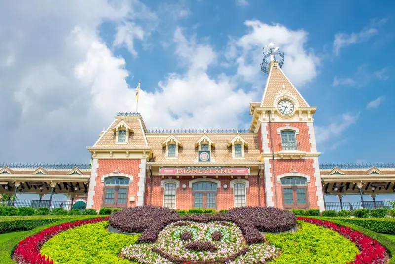 Disneyland entrance gate with Victorian architecture, clock tower, and colorful Mickey Mouse flower garden under blue sky