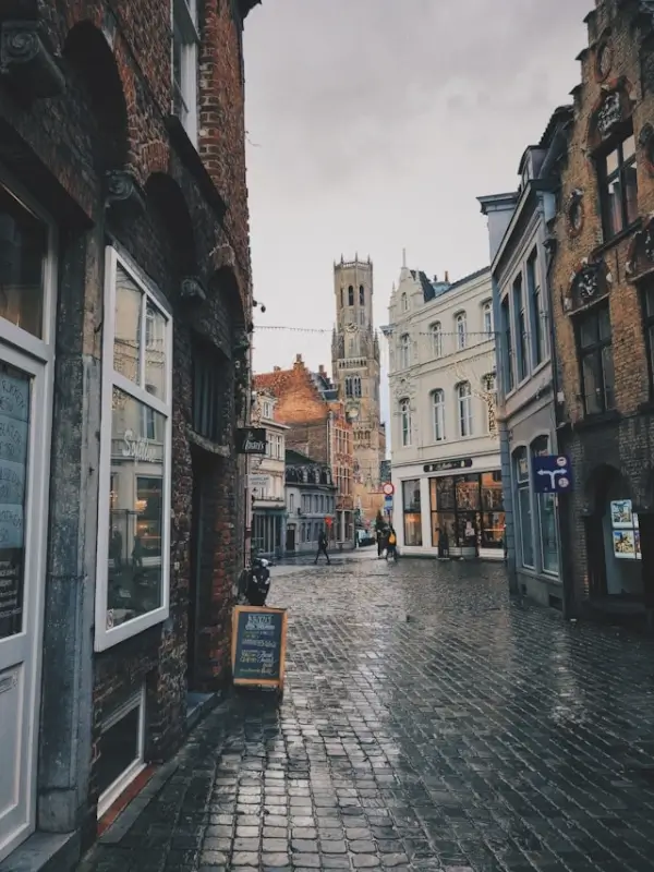 Cobblestone street in medieval Bruges with historic brick buildings, shops, and the famous Belfry tower in the background