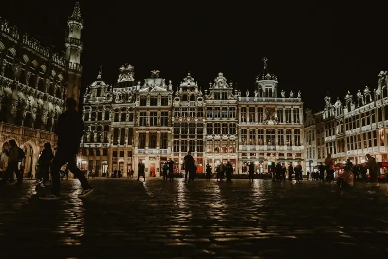 Brussels Grand Place at night showing illuminated Gothic guildhalls and Town Hall with tourists walking across the cobblestone square