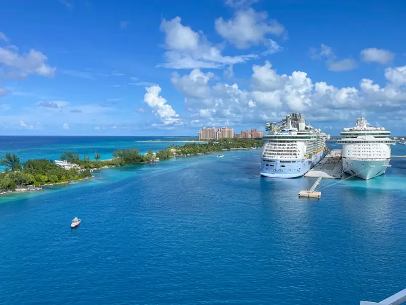 Two large cruise ships docked at a Caribbean port with turquoise waters, tropical islands, and resort buildings under blue skies