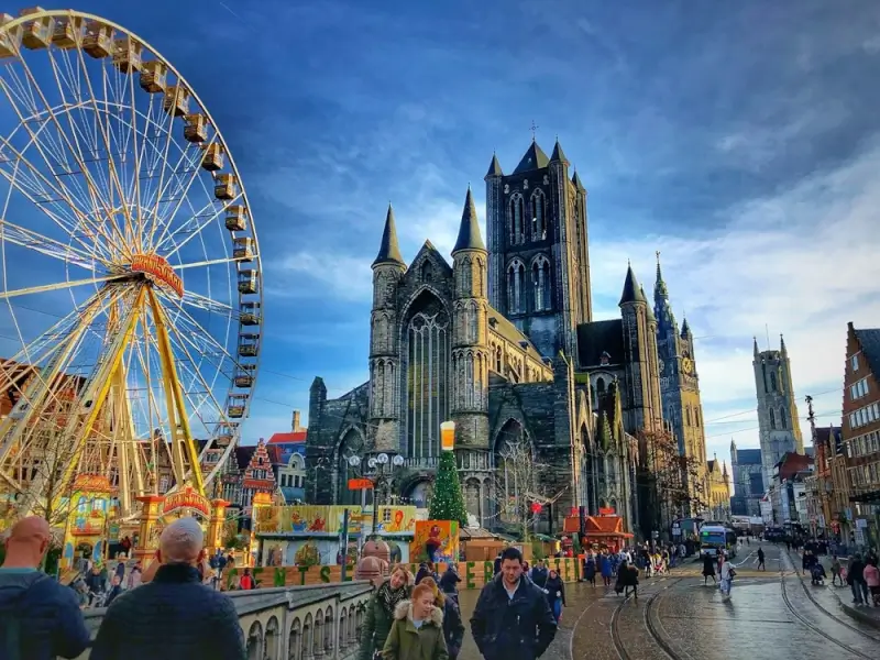 Ghent's Saint Nicholas Church and Belfry tower with a Ferris wheel and festival market in the foreground showing crowds of visitors