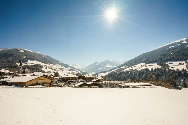 Picturesque Austrian Alpine village in Alpbach with traditional wooden chalets and church spire under bright winter sun, surrounded by snow-covered mountains and forested valleys