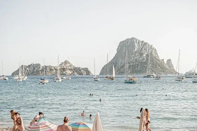Beachgoers relaxing at Ibiza beach with Es Vedrà rock formation and sailboats in Mediterranean waters
