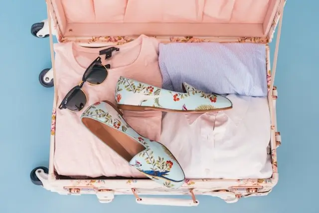 Open pink suitcase with comfortable floral flat shoes, sunglasses, and neutral cruise clothing packed for luxury vacation travel