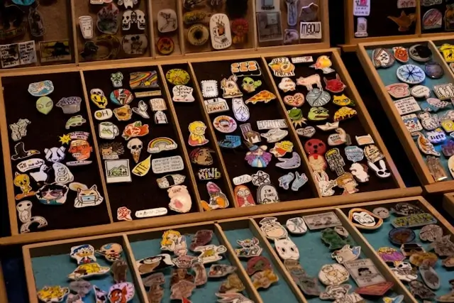 Wooden display case with multiple compartments showcasing a large collection of colorful enamel pins and patches. The collection includes various designs featuring skulls, cartoon characters, food items, animals, text phrases, and pop culture references, organized across different sections of the display case