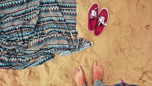 Beach outfit flatlay with patterned cover-up, red sneakers and feet on sand in Ibiza