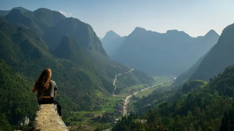 Traveler overlooking Ha Giang Loop valley from mountain viewpoint with winding road through limestone peaks and rice terraces in northern Vietnam
