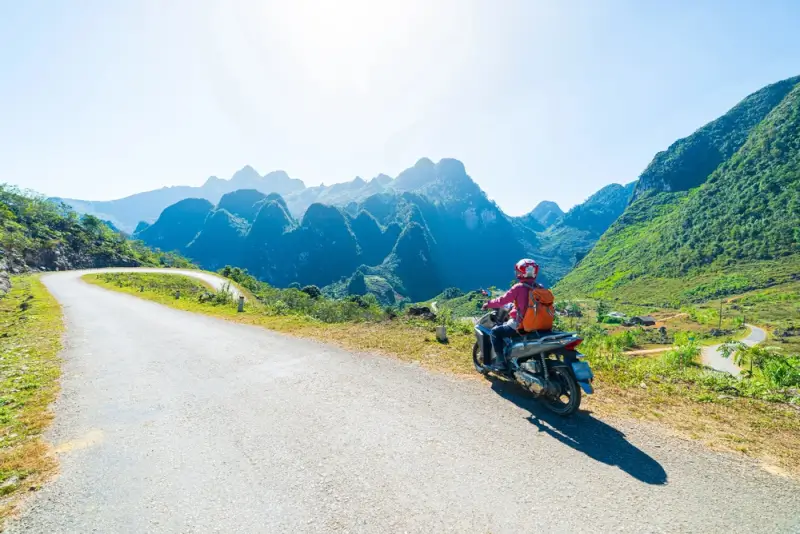 Solo motorbike traveler with backpack riding Ha Giang Loop with stunning limestone karst mountains and clear blue sky in northern Vietnam