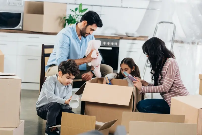 A family packing household items into cardboard boxes together in their kitchen while preparing for a relocation overseas.