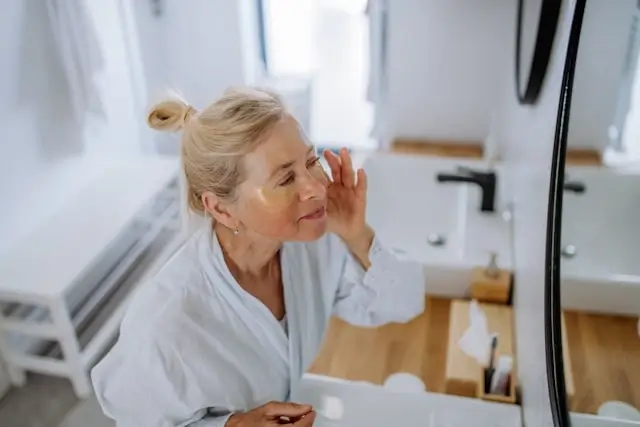 Mature woman in white bathrobe applying skincare product while looking in bathroom mirror