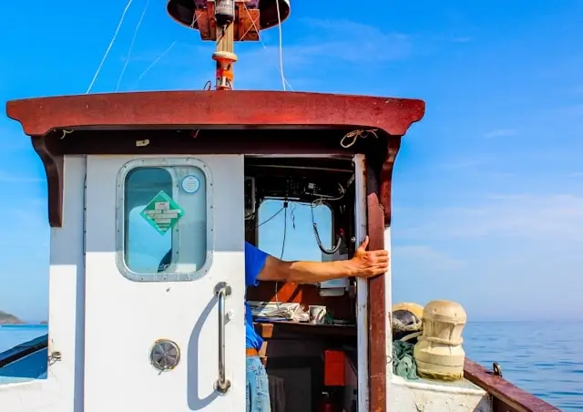 Fishing boat crew member in blue shirt operating vessel from pilothouse on calm ocean waters
