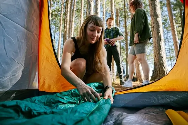 Young woman setting up camping tent in forest with friends, demonstrating essential outdoor gear for weekend adventures
