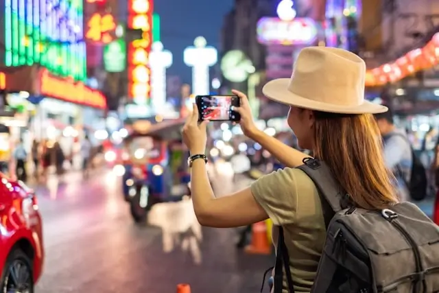 Female traveler with backpack taking photos in busy city street with colorful neon lights at night
