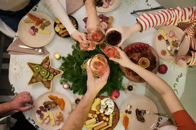 Top view of friends raising glasses for a toast over a festive Christmas dinner table decorated with greenery, cheese platters, fruits, and holiday snacks.