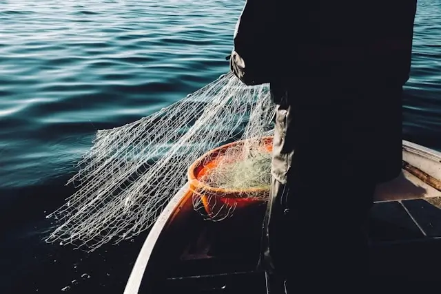 Fisherman in dark apparel handling fishing net with fresh catch on boat deck over calm blue waters