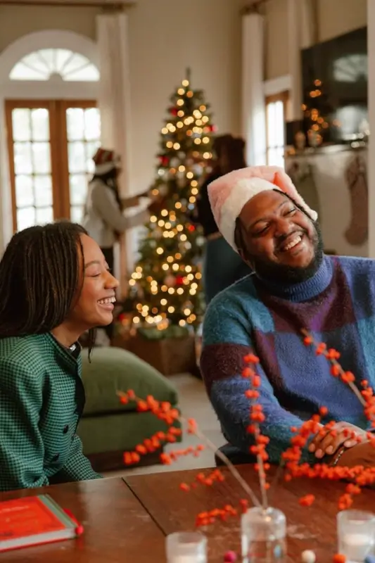 Two people smiling and chatting at a cozy Christmas gathering, with a decorated tree and stockings in the background.