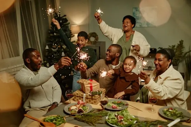Family celebrating around a Christmas dinner table, smiling and holding sparklers with wrapped gifts and holiday decorations nearby.