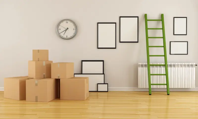 Cardboard moving boxes stacked against a wall in an empty room with picture frames and a ladder, representing packing belongings for an international relocation.