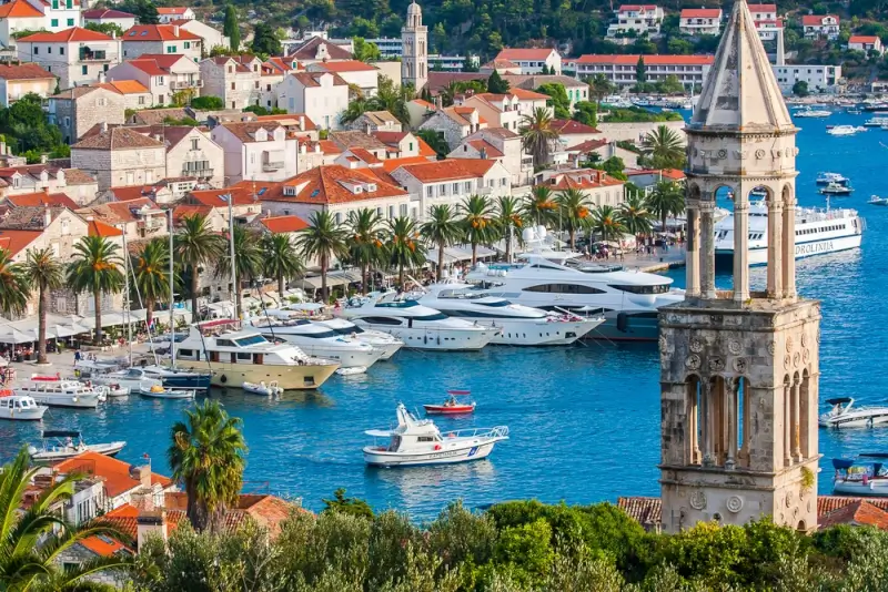 Luxury yachts anchored in Hvar harbor beside historic bell tower with palm-lined waterfront and red-roofed buildings