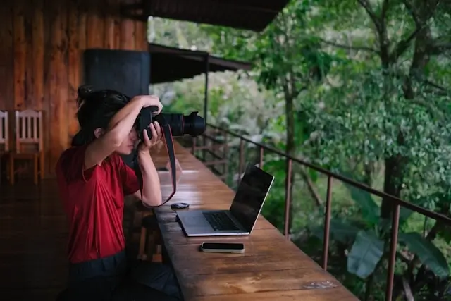 Woman photographer taking pictures with laptop on wooden balcony in nature, creating content for travel blog