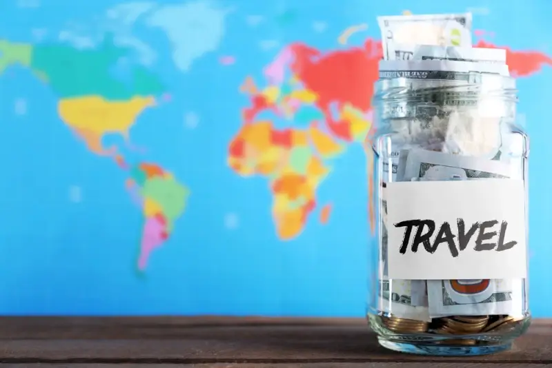 Travel savings jar filled with money and coins against world map background for vacation budget