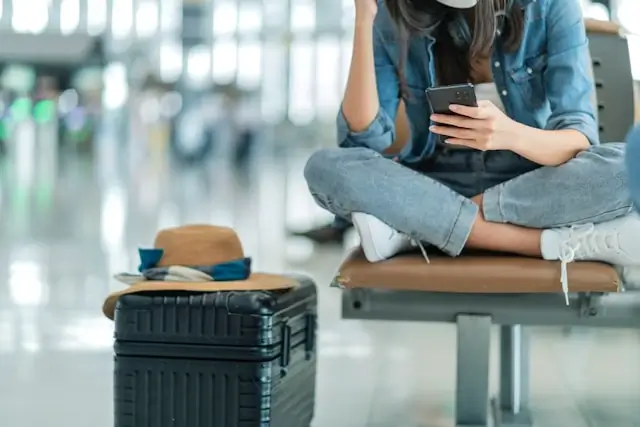 Traveler sitting on airport bench with luggage and phone during flight delay covered by travel insurance