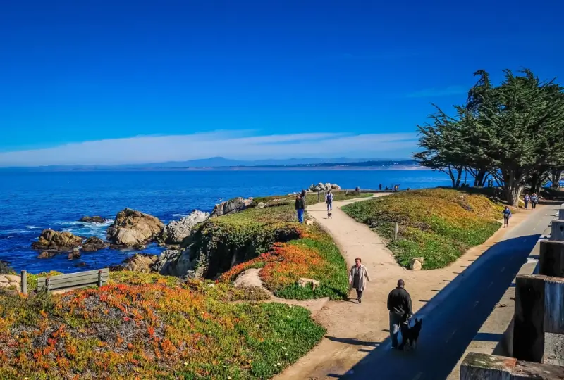 Coastal walking trail along the Monterey Peninsula with ocean views, rocky shoreline, and people strolling beside the Pacific Ocean on a clear day.