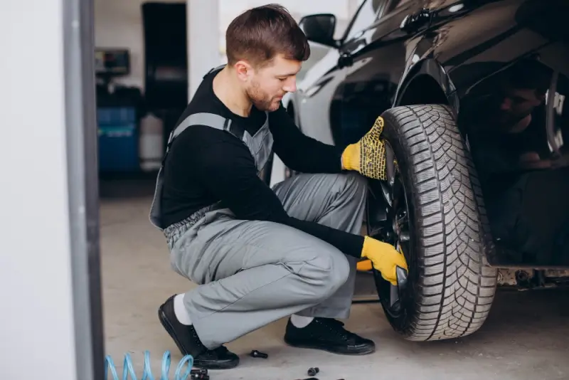 A mechanic kneeling beside a lifted car while removing a front wheel, wearing yellow gloves and work overalls inside an auto repair shop.