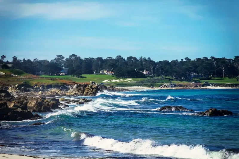 Waves crashing along the rocky shoreline at Pebble Beach, with a coastal golf course and pine trees overlooking the Pacific Ocean.