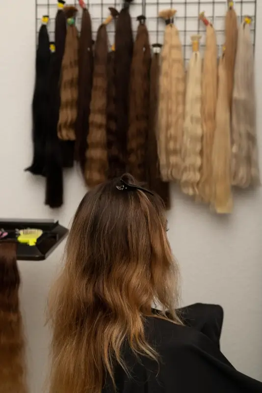 Woman seated in a beauty salon chair with natural hair visible, while multiple bundles of human hair extensions in different lengths and shades hang on a display rack behind her.