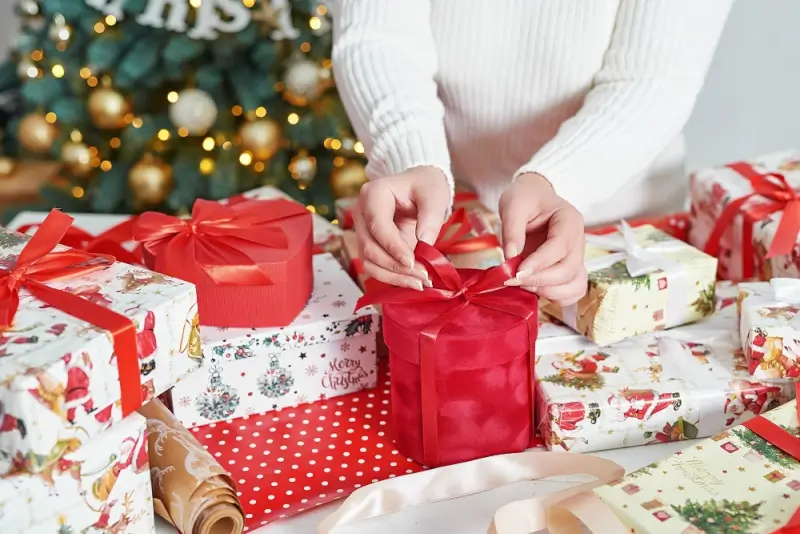 Woman wrapping holiday presents with red ribbon bows near decorated christmas tree with festive gift paper