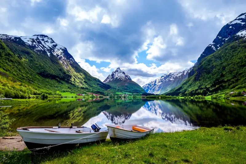 Two boats resting on grassy shore beside mirror-like fjord reflecting snow-capped mountains and green valley in Norwegian wilderness