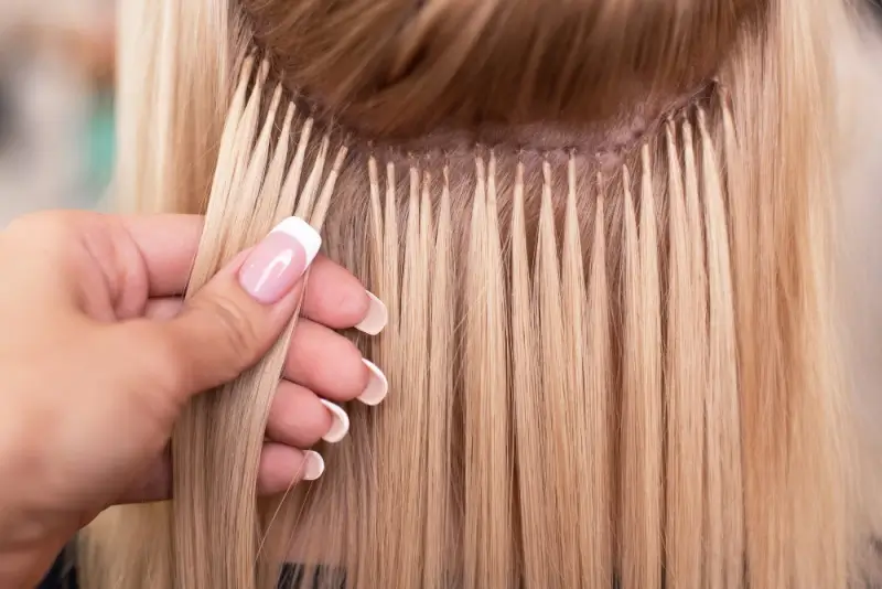 Close-up of blonde hair extensions being applied section by section, showing precise attachment near the roots for a seamless, natural blend.