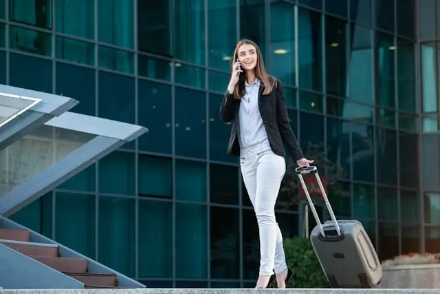 Professional woman in business attire walking with rolling suitcase while on phone call outside modern office building