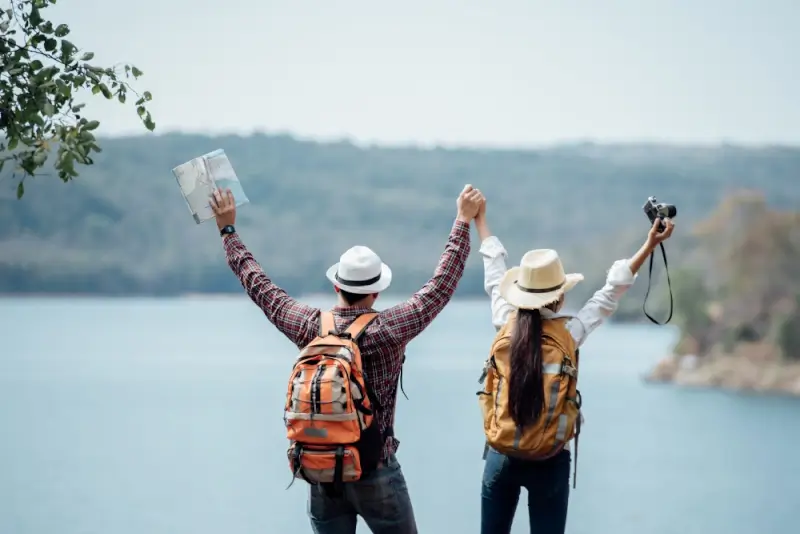 Couple with backpacks and sun hats celebrating at scenic lakeside viewpoint holding map and camera while traveling together