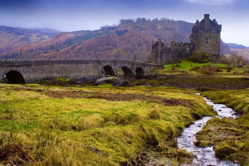 Historic stone castle beside an arched bridge in the Scottish Highlands, surrounded by rolling hills, grassy fields, and a winding stream.