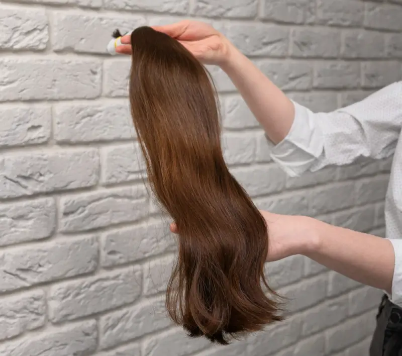 Hand holding a single bundle of long brown human hair extension against a textured wall, showing hair length, smooth texture, and natural shine.