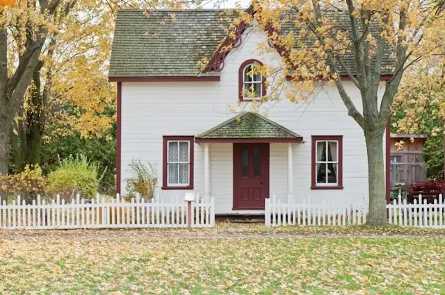 Charming white cottage with red trim and picket fence surrounded by autumn trees, illustrating the warmth of making a house a home