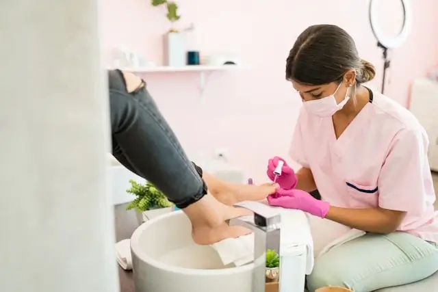 Nail technician in pink gloves performing meticulous pedicure prep work in clean Dubai salon with client's foot elevated for precision treatment