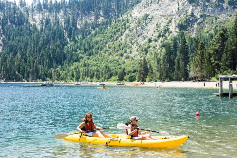 Two people kayaking on a clear mountain lake surrounded by pine forests and rocky hills on a sunny day in California.
