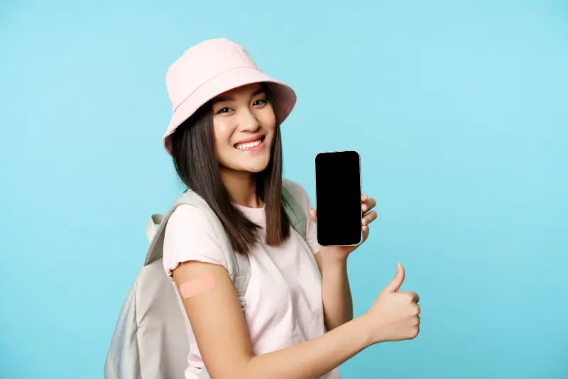 Smiling female traveler in pink bucket hat holding smartphone with emergency contact numbers giving thumbs up