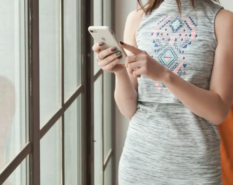 Person standing by a window while using a smartphone, representing online dating, messaging, and making connections through mobile dating platforms.