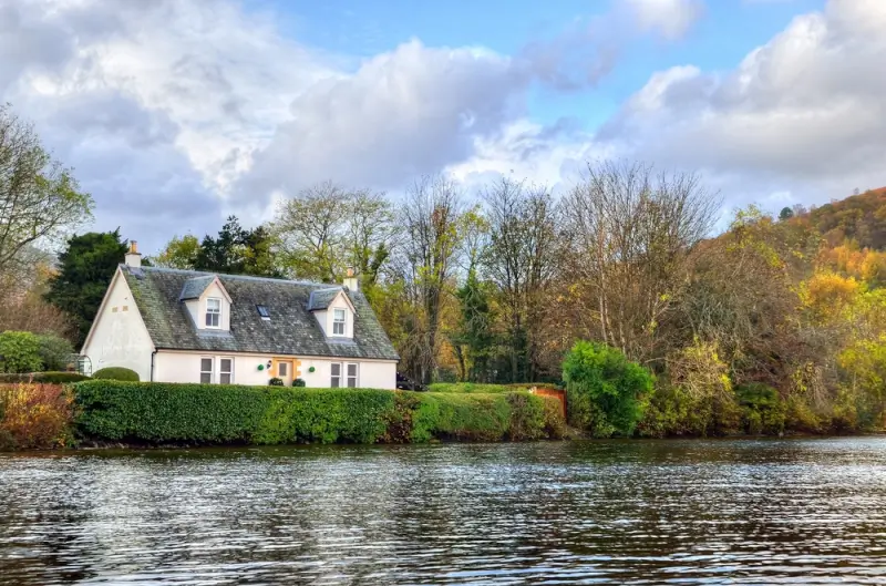 Charming cottage set beside a calm loch in Scotland, surrounded by autumn trees and peaceful waterside scenery.
