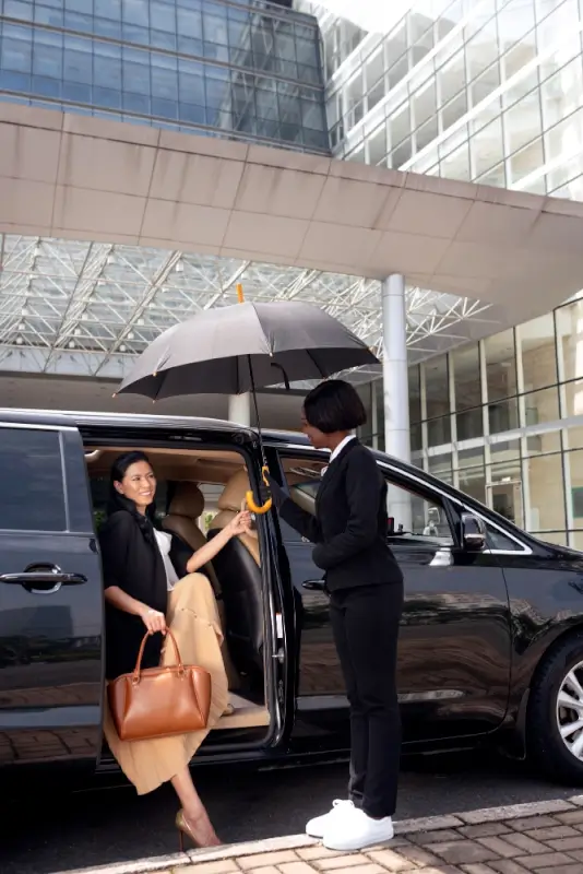 Professional chauffeur holding umbrella for female passenger exiting luxury vehicle at modern airport terminal entrance