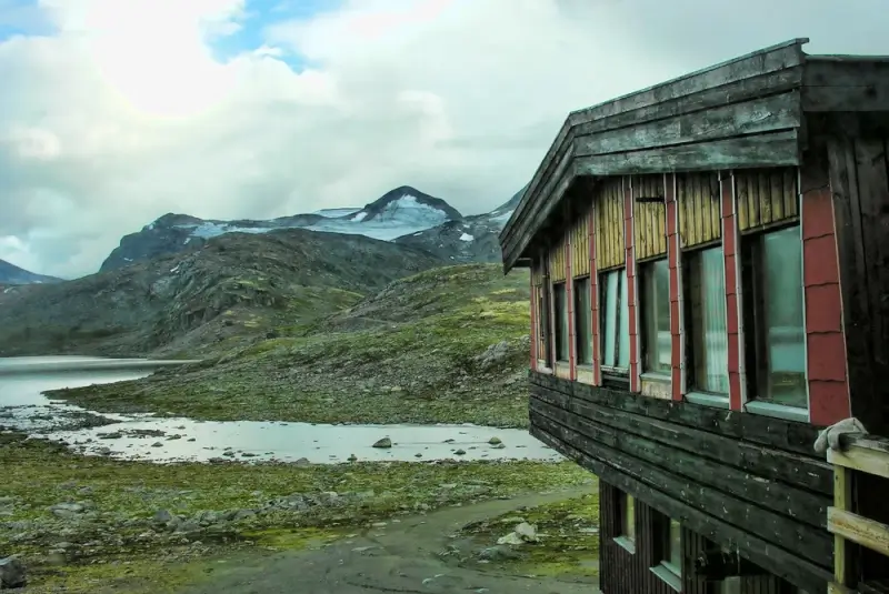 Rustic DNT hut with colorful shuttered windows overlooking alpine lake and glacier-capped mountain in Jotunheimen National Park