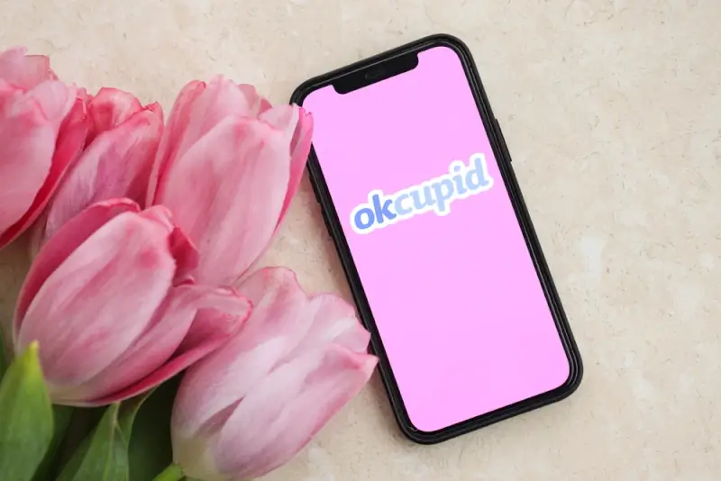 Smartphone displaying the OkCupid dating app logo placed beside pink tulips, symbolizing modern romance and compatibility-focused online dating.