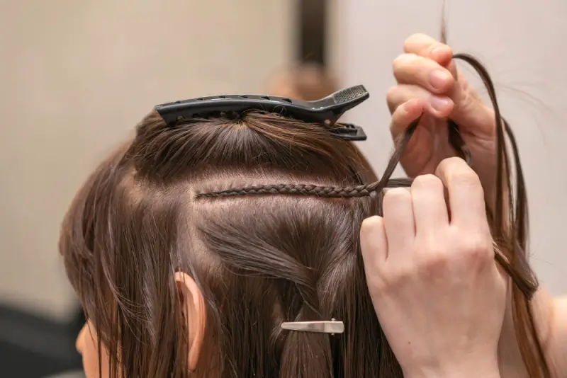Stylist braiding natural hair close to the scalp during a sew-in hair extension installation, with sectioned hair clipped neatly for precision work.
