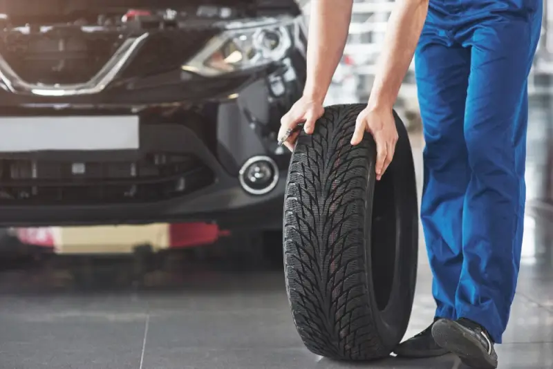 A mechanic in blue work pants rolling a car tire across the shop floor with the hood of a black vehicle open in the background.